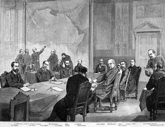 Homens sentados em torno de uma grande mesa; no fundo da sala, um mapa do continente africano, alvo do neocolonialismo.
